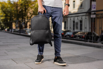 A black leather backpack holding male hand in city - obrazy, fototapety, plakaty