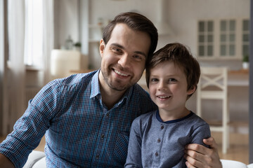 Portrait of happy millennial dad and preschooler son kid looking at camera, speaking, speaking on video call. Father and child sitting on couch together, looking at webcam with happy smile, hugging