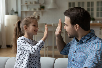 Happy proud dad and daughter giving high five, having fun together, enjoying activity, sitting on couch. Smiling father and girl playing at home, making deal. Daddy giving praise, approval, support