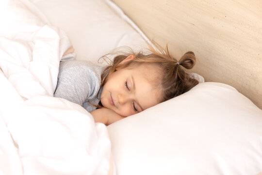 Little adorable child girl in grey pajamas closed eyes lying in bed sleeping on comfortable pillow and under white fluffy cotton blanket, good night sweet dreams