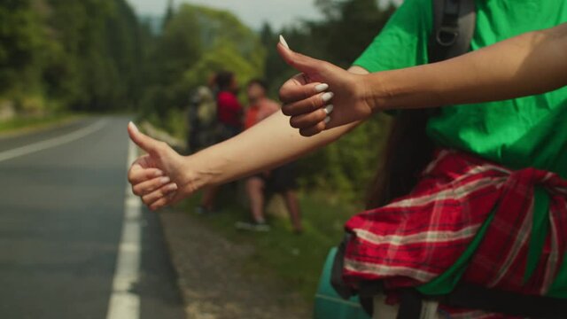 Close-up of multiracial female hands with perfect manicured nails signing thumbs up while hitchhiking at mountain road during adventure trek, with blurry diverse friends relaxing in background.