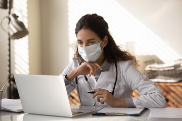 Young female general practitioner in medical face mask working at laptop alone, making video call, reading electronic medical records. Doctor giving online consultation to patients from covid hospital