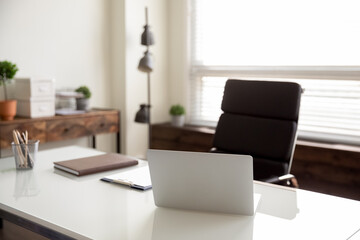 Businessperson empty office workplace with no people, comfortable armchair for work at white desk with open laptop placed by window in pale colored room. Modern business leader table with computer