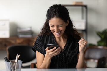 Happy millennial Latin woman in casual excited with good news, using smartphone, looking at screen,...