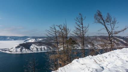 Fototapeta na wymiar The blue ice-free Angara River flows between the snow-covered banks. Trunks and bare branches of trees against the background of the sky and mountains. A section of the frozen Lake Baikal is visible.