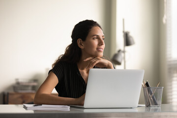 Smiling thoughtful female employee working at laptop from home, looking away, planning tasks,...