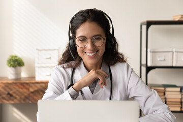 Happy friendly female physician in headphones chatting to patient on video call, giving help,...