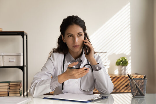 Serious female doctor giving telephone consultation, talking to patient on cellphone, making phone call from clinic office. General practitioner working on healthcare hotline, answering questions - Powered by Adobe