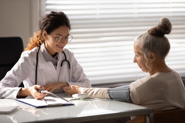 Smiling friendly doctor checking blood pressure of older female patient at appointment in office,...