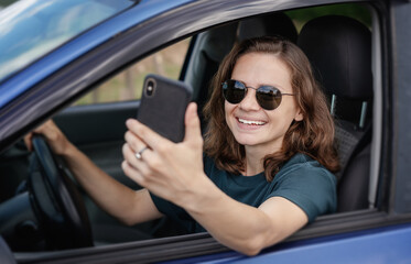 Fototapeta na wymiar Beautiful happy young woman driver sitting in car wearing sunglasses using smartphone