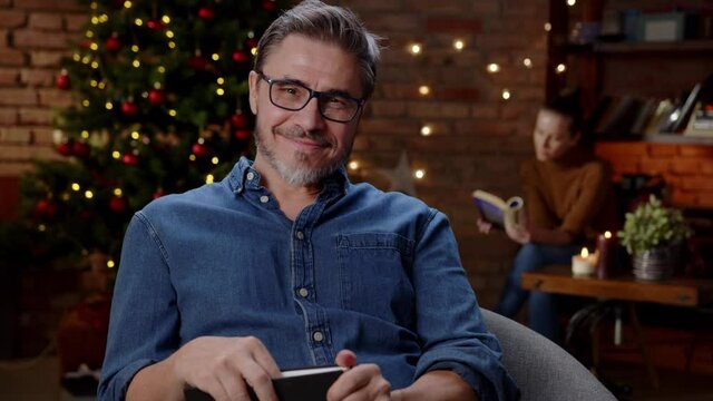 Couple reading books at home at Christmas in winter. Happy older white man in glasses, smiling while reading, dark room with Christmas tree.
