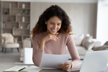 Happy young attractive hispanic woman reading correspondence paper letter with good news, getting bank mortgage last payment notification or taxes refund document, sitting alone at table at home.