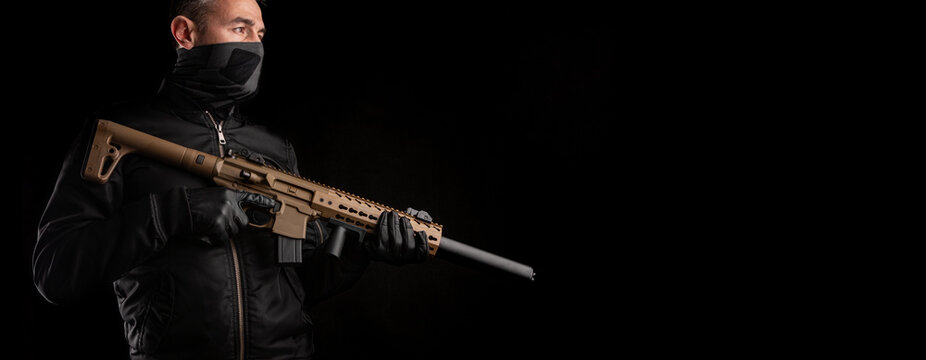 A man in dark clothes holds an automatic carbine in his hands. The face is covered with a mask, hands are in gloves. A figure with a weapon on a dark back