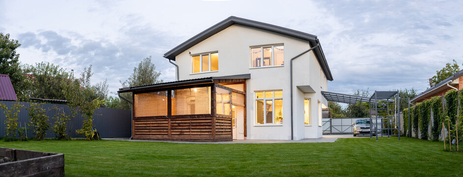 Exterior view of modern white house with courtyard, patio area, green grass lawn, garden and a car in the evening
