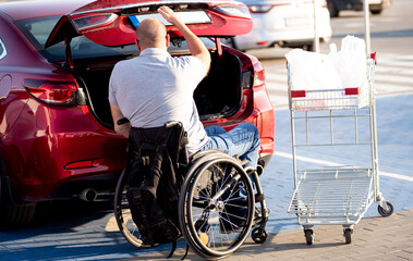 Fototapeta na wymiar Adult disabled man in a wheelchair puts purchases in the trunk of a car in a supermarket parking lot