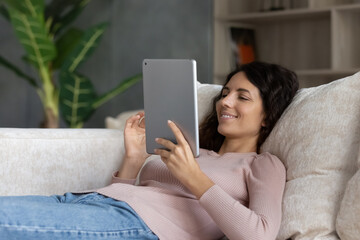 Happy lazy millennial hispanic woman using digital touchpad software applications, web surfing information, choosing goods shopping in internet store, communicating distantly lying on cozy sofa.