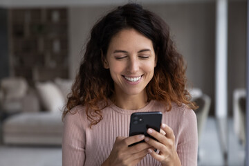 Smiling attractive young hispanic woman reading pleasant message on cellphone, communicating distantly with friends in social networks or using e-dating application, shopping online or web surfing.