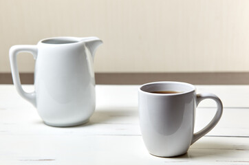 White cup with coffee and milk jug on a white wooden background, closeup. Energy breakfast, morning routine concept