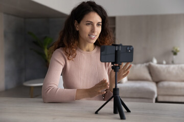 Smiling millennial hispanic woman talking recording video on cellphone web camera standing on...
