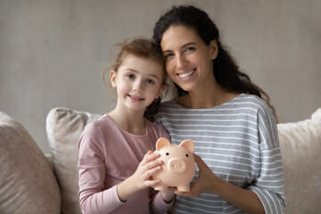 Portrait of smiling young hispanic mother cuddling little cute kid daughter, holding piggybank in hands, learning planning family budget, saving for future investment or insurance, financial literacy.
