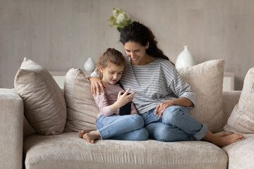 Fototapeta premium Happy small kid girl using telephone applications, playing mobile games or spending time online resting on comfortable sofa with affectionate caring young mother, modern technology addiction concept.