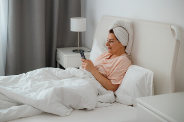 Happy young caucasian woman relax on comfortable bed at home texting messaging on smartphone, smiling girl use cellphone, browse wireless internet on gadget. Concept of shopping online from home