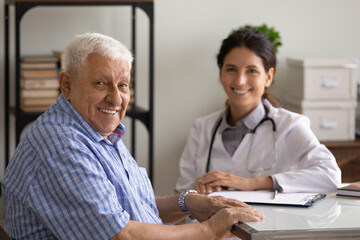 Portrait of happy elder 80s man and young female doctor at appointment. Senior patient visiting therapist in office, looking at camera, smiling, Geriatric healthcare, elderly medic care concept