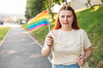 Beautiful red lesbian woman with LGBT rainbow flag at sunset on green nature background. Happiness, freedom and love concept for same sex couples.