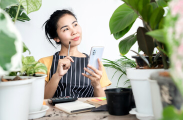 Asian woman selling plants online to start up new business , successful female shop owner sitting at home using mobile phone and writing orders
