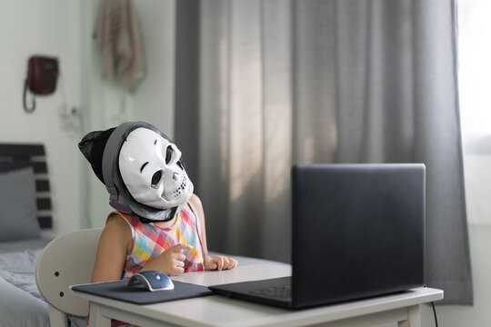 Child wondering tilt head or kid girl learning on computer notebook and wearing ghost skull mask with headset for video call study online to thinking or people learn from home by laptop on halloween