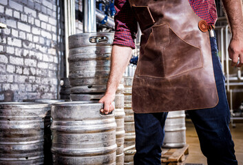 Obraz na płótnie Canvas Young male brewer in leather apron holds barrel with craft beer at modern brewery factory