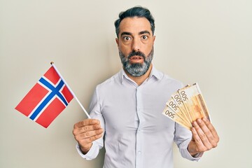 Middle age man with beard and grey hair holding norway flag and krone banknotes in shock face, looking skeptical and sarcastic, surprised with open mouth