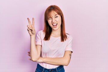 Redhead young woman wearing casual pink t shirt smiling with happy face winking at the camera doing victory sign. number two.