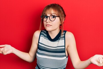 Redhead young woman wearing casual clothes and glasses clueless and confused expression with arms...