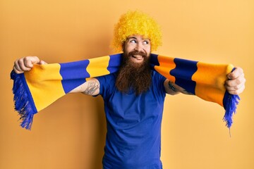 Redhead man with long beard football hooligan cheering game wearing funny wig smiling looking to...