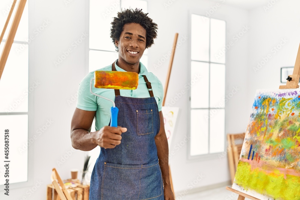 Poster Young african american artist man holding paint roller standing at art studio.