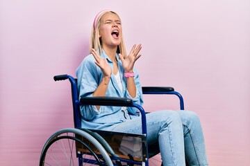Beautiful blonde woman sitting on wheelchair celebrating mad and crazy for success with arms raised...