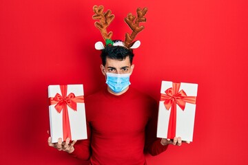 Young hispanic man wearing cute christmas reindeer horns and medical mask holding gifts depressed and worry for distress, crying angry and afraid. sad expression.