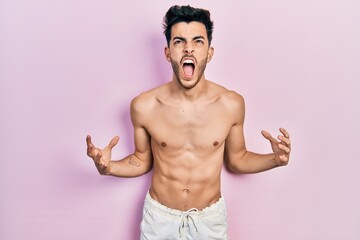 Fototapeta premium Young hispanic man wearing swimwear shirtless crazy and mad shouting and yelling with aggressive expression and arms raised. frustration concept.