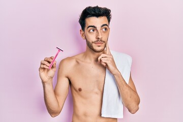 Fototapeta premium Young hispanic man standing shirtless holding razor serious face thinking about question with hand on chin, thoughtful about confusing idea