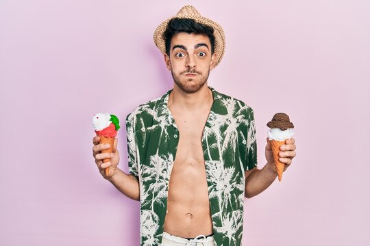 Young hispanic man wearing summer style holding ice cream puffing cheeks with funny face. mouth inflated with air, catching air.