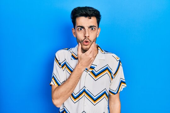 Young hispanic man wearing casual clothes looking fascinated with disbelief, surprise and amazed expression with hands on chin