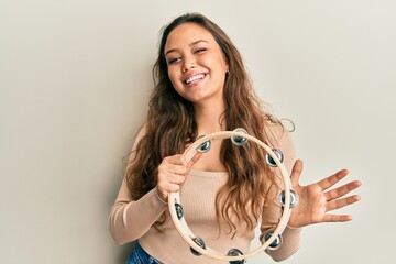 Young hispanic girl playing tambourine looking positive and happy standing and smiling with a confident smile showing teeth - obrazy, fototapety, plakaty
