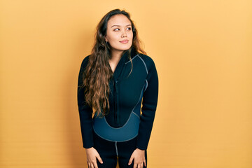 Young hispanic girl wearing diver neoprene uniform smiling looking to the side and staring away...