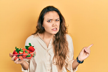 Young hispanic girl holding strawberries pointing aside worried and nervous with forefinger,...