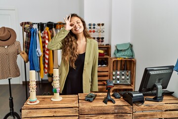 Beautiful hispanic woman working at fashion shop doing ok gesture with hand smiling, eye looking through fingers with happy face.