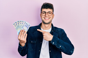 Young hispanic man holding 100 romanian leu banknotes smiling happy pointing with hand and finger