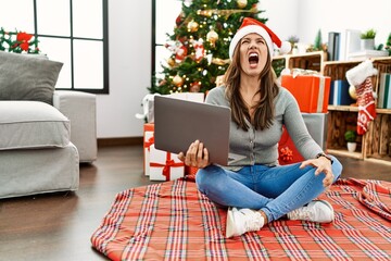 Obraz na płótnie Canvas Young latin woman using laptop sitting by christmas tree angry and mad screaming frustrated and furious, shouting with anger. rage and aggressive concept.