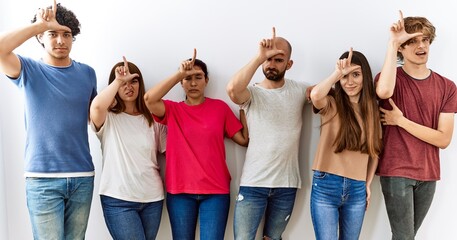 Group of young friends standing together over isolated background making fun of people with fingers on forehead doing loser gesture mocking and insulting.