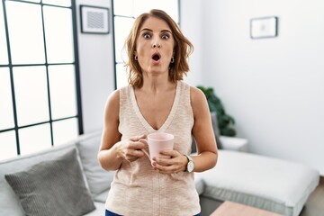 Middle age woman drinking a cup coffee at home in shock face, looking skeptical and sarcastic, surprised with open mouth